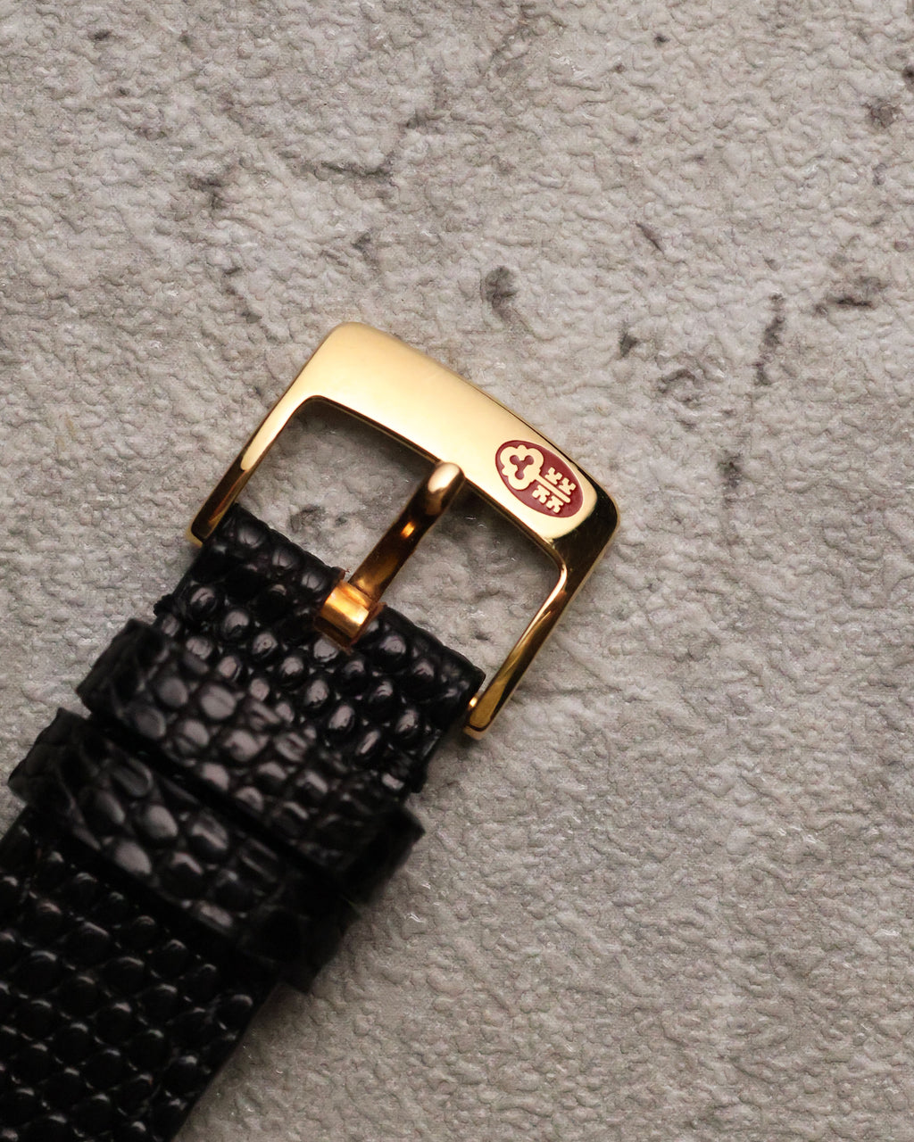 Close-up of the original 18k yellow gold Corum buckle, engraved with the Corum key logo, attached to the black textured leather strap.