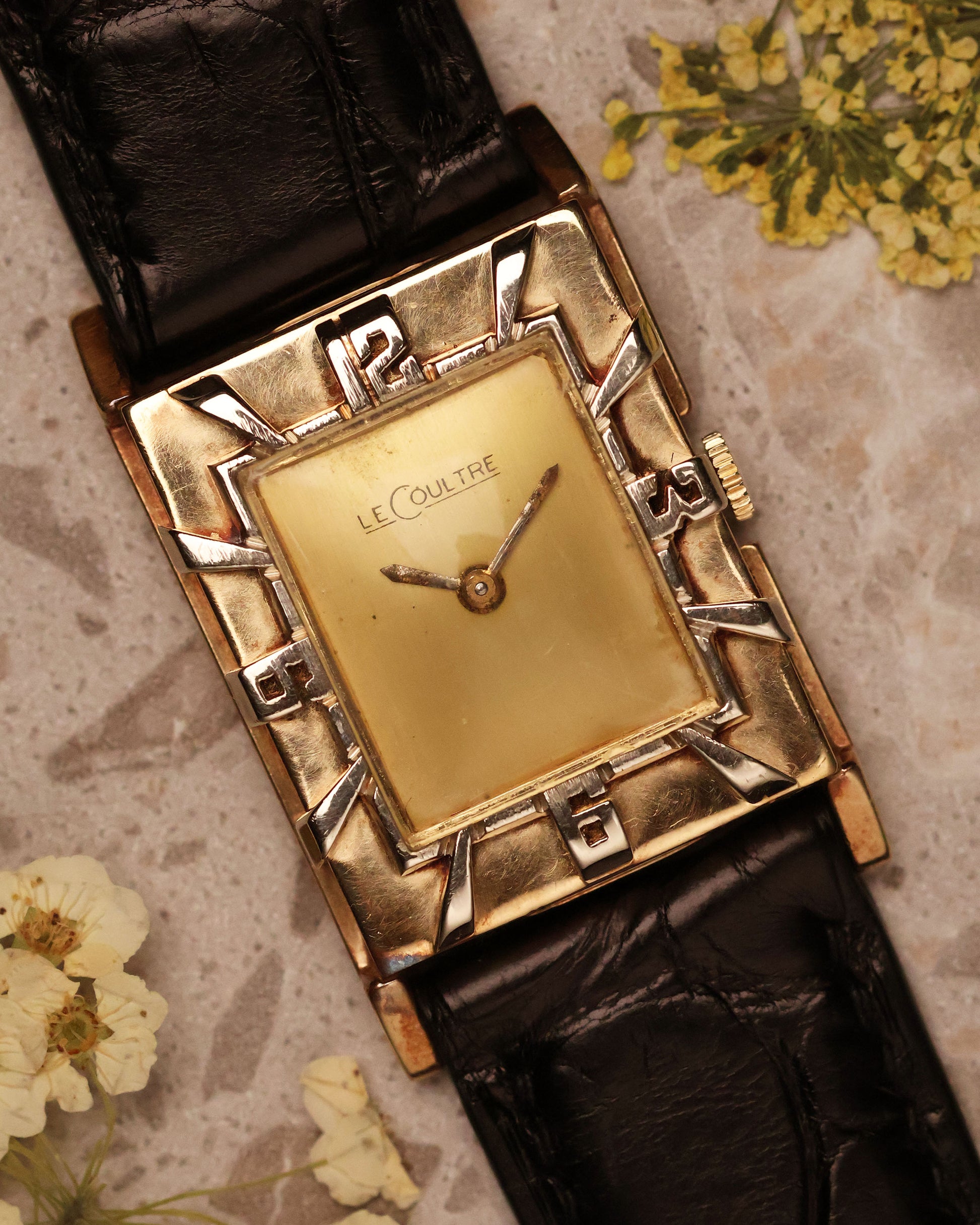 Top-down view of the LeCoultre 14K gold vintage watch with Art Deco numerals bezel, gold dial, and black leather strap on a marble surface.