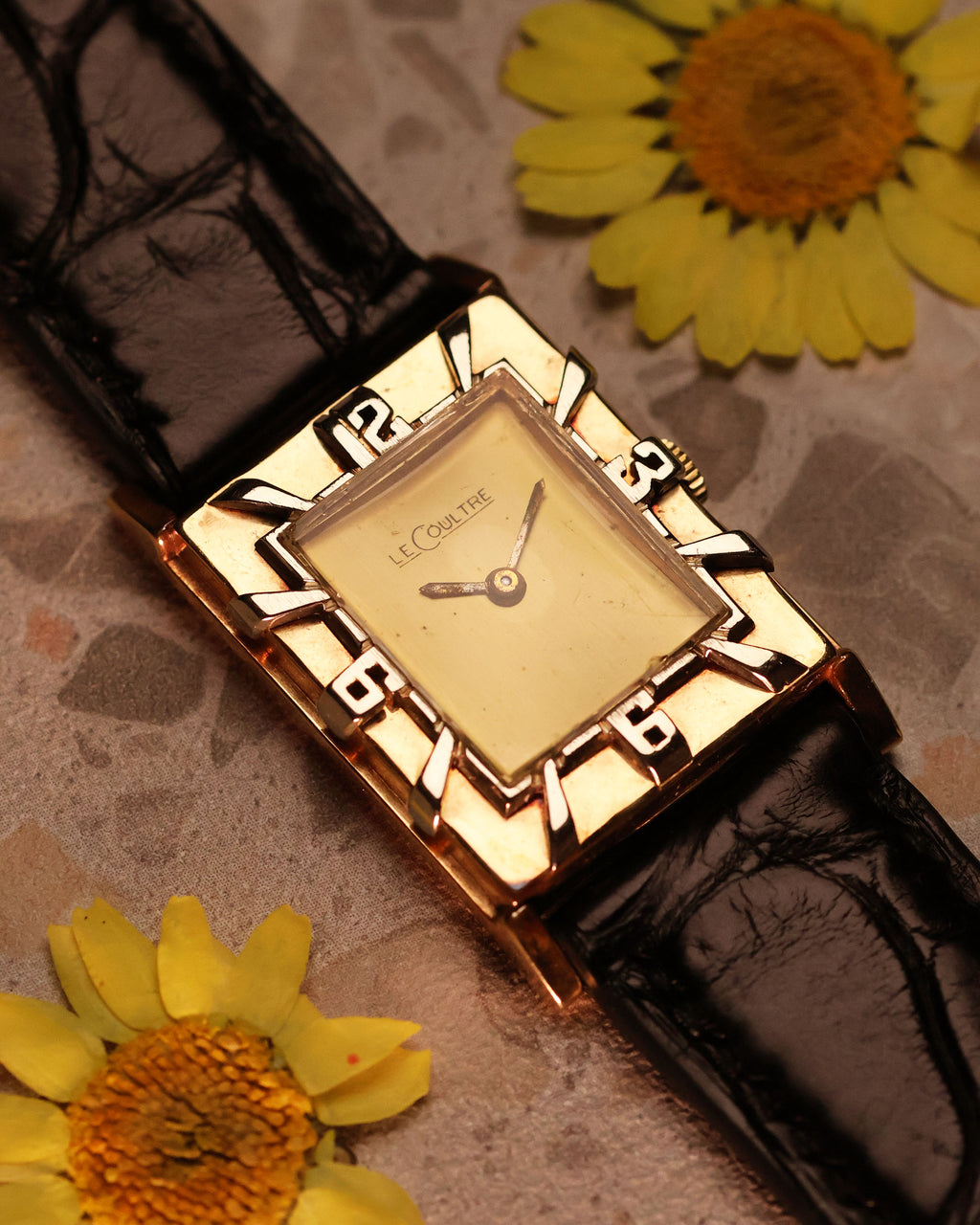 LeCoultre 14K gold tank watch placed on marble with yellow flowers, showcasing the interplay of gold tones and Art Deco numerals bezel.
