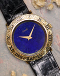 Close-up of a Piaget Ref. 9118 watch with a deep blue lapis lazuli dial and an ornate engraved 18K yellow gold bezel on a black leather strap.