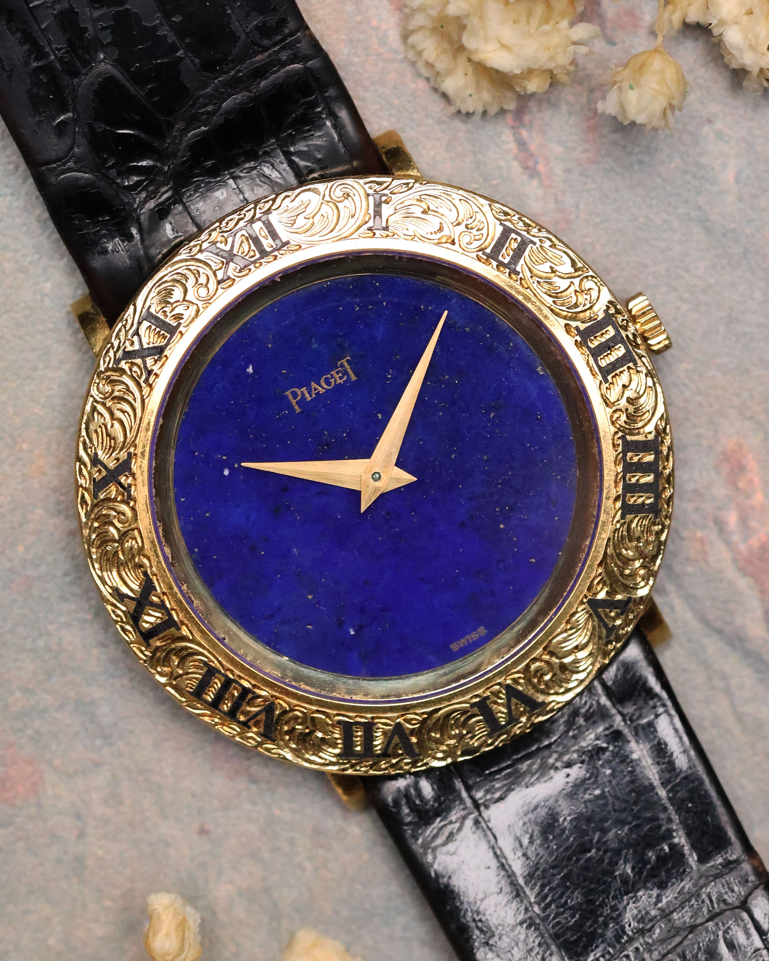 Close-up of a Piaget Ref. 9118 watch with a deep blue lapis lazuli dial and an ornate engraved 18K yellow gold bezel on a black leather strap.