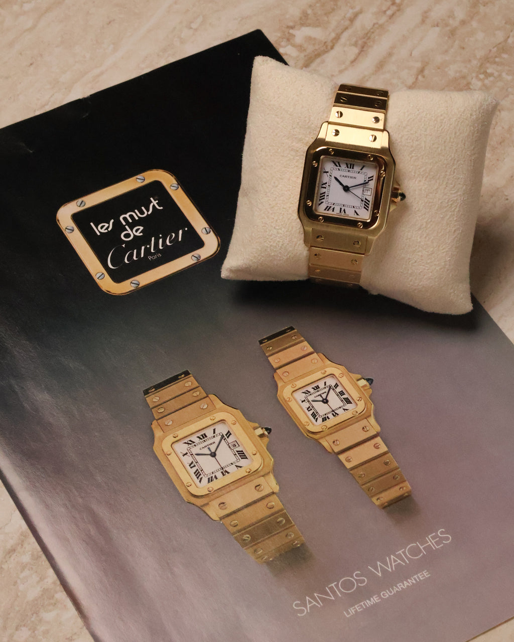 Cartier Santos Carrée Ref. 2960 in 18K yellow gold resting on a cushion, photographed beside a vintage ‘Les Must de Cartier’ Santos brochure featuring two gold Santos models.