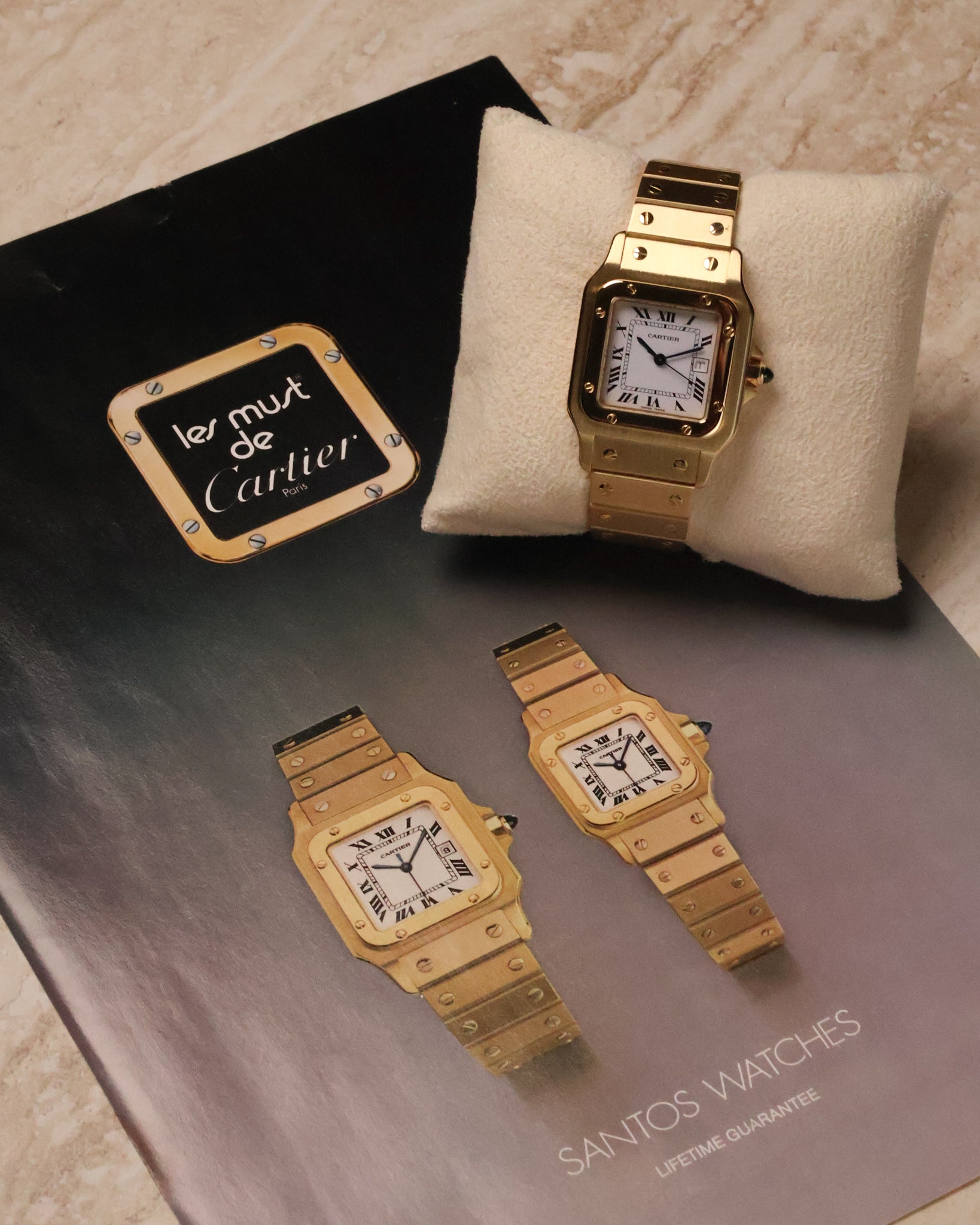 Cartier Santos Carrée Ref. 2960 in 18K yellow gold resting on a cushion, photographed beside a vintage ‘Les Must de Cartier’ Santos brochure featuring two gold Santos models.