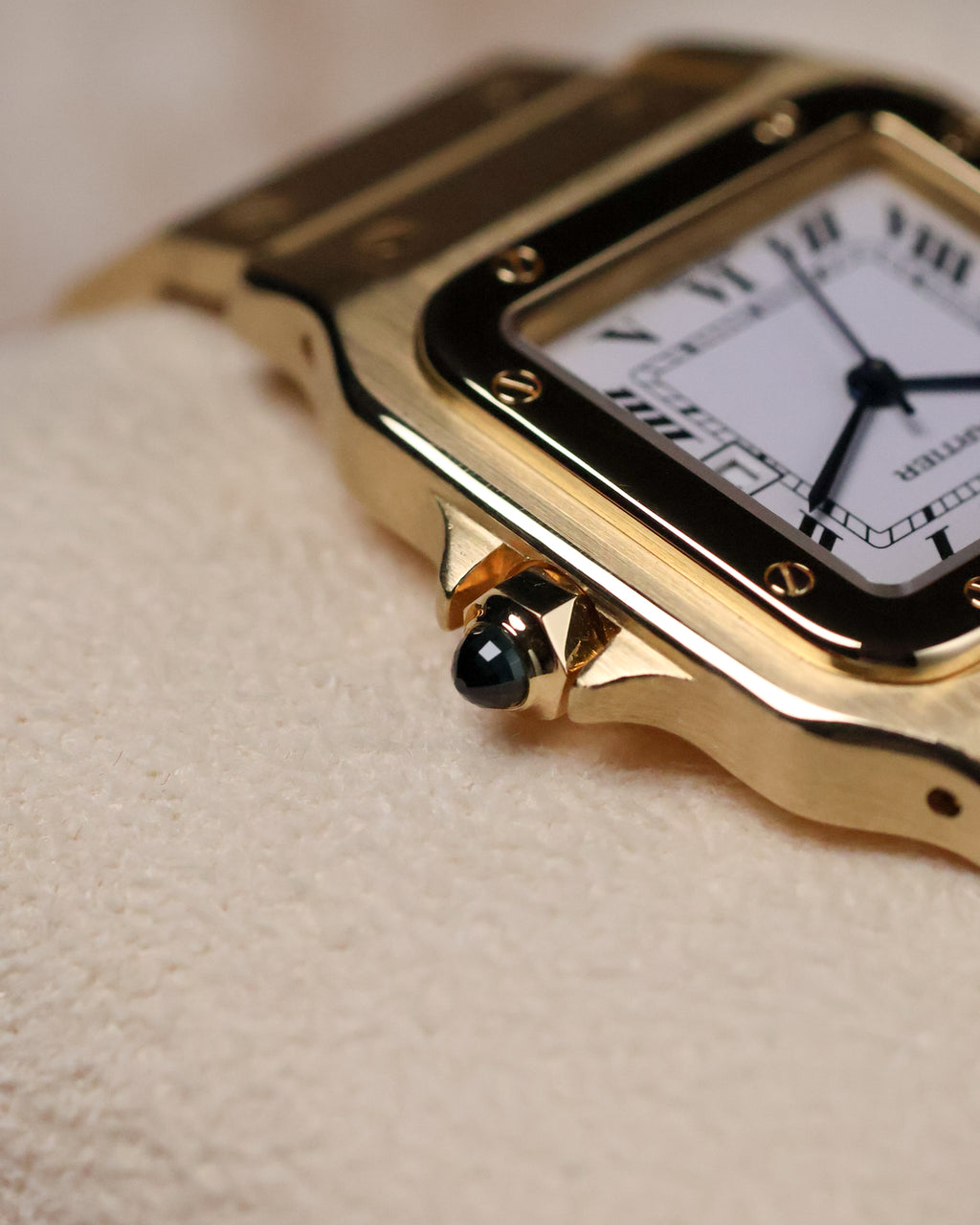 Macro shot of the Cartier Santos Carrée crown featuring the blue cabochon, with the gold case and bezel screws visible along the dial edge.