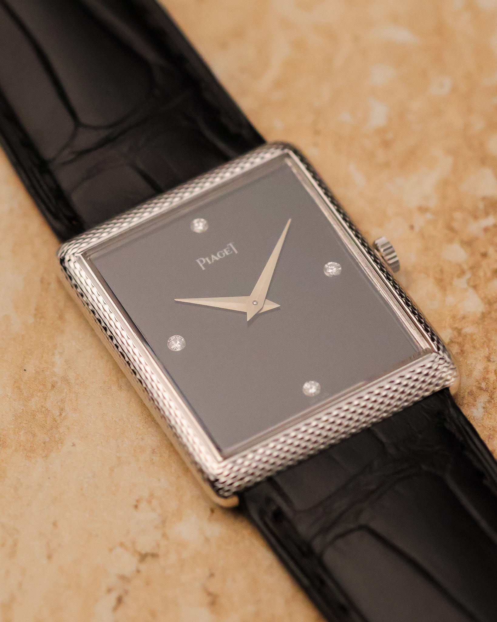 Three-quarter angled view of Piaget 9152 highlighting onyx dial, diamond markers and woven case detailing against light stone background.